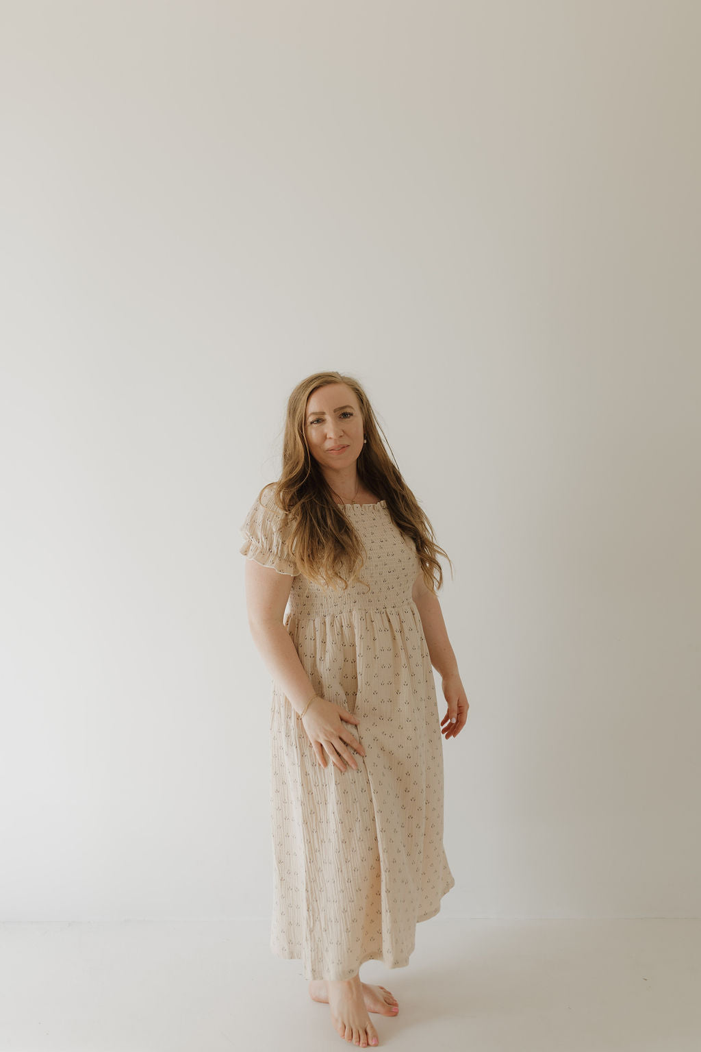 A woman with long light brown hair stands barefoot in the Women's Short Sleeve Muslin Dress | Flora by forever french baby, an ankle-length, light-colored cotton dress, against a plain background, calmly looking at the camera.
