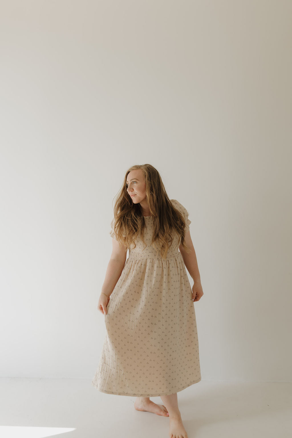 A barefoot woman with long hair stands against a light background, looking left and gently holding her Women's Short Sleeve Muslin Dress | Flora by forever french baby.