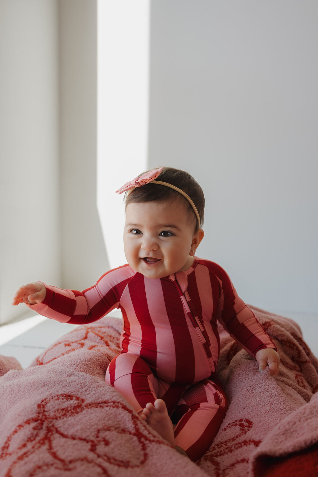 A smiling baby in Minty Cherries Bamboo Zip Pajamas | Iconic Stripe and a pink bow headband sits on a pink blanket in a sunlit room.