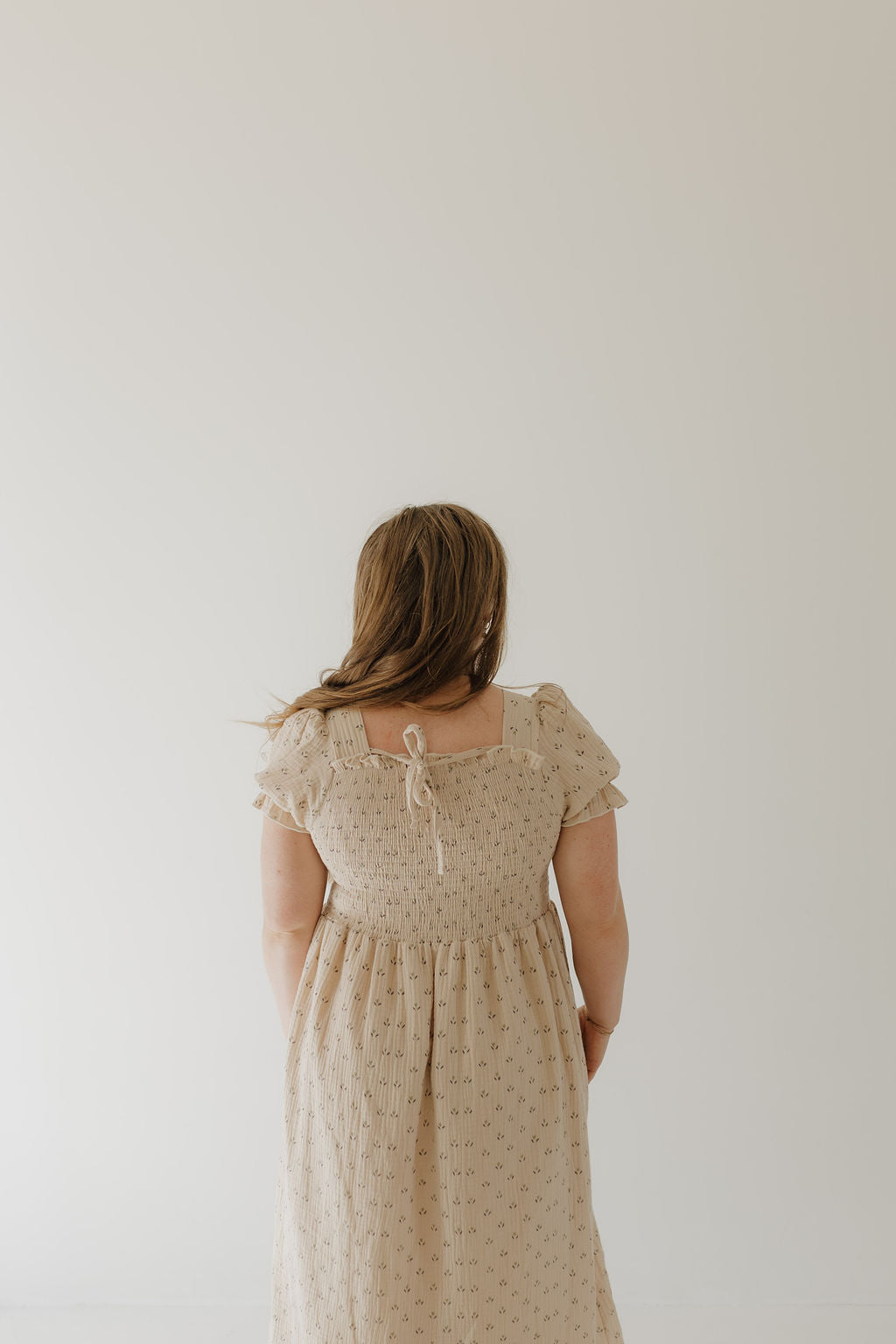 A woman with long brown hair stands with her back to the camera in a beige Women's Short Sleeve Muslin Dress | Flora by forever french baby, featuring lightweight cotton, a smocked back, and a bow at the neckline.
