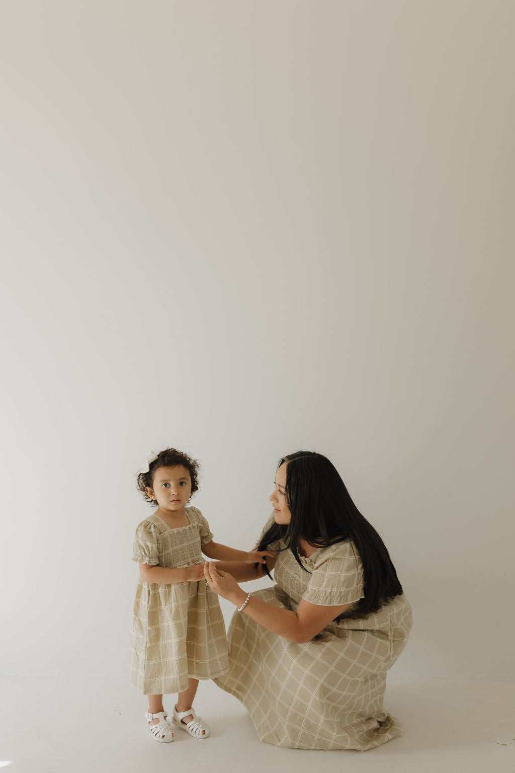 A woman kneels beside a young girl, both in matching forever french baby Toddler Muslin Dress | Linen Grid outfits, featuring 100% cotton beige plaid, in a minimal, light-filled setting with a plain off-white background.