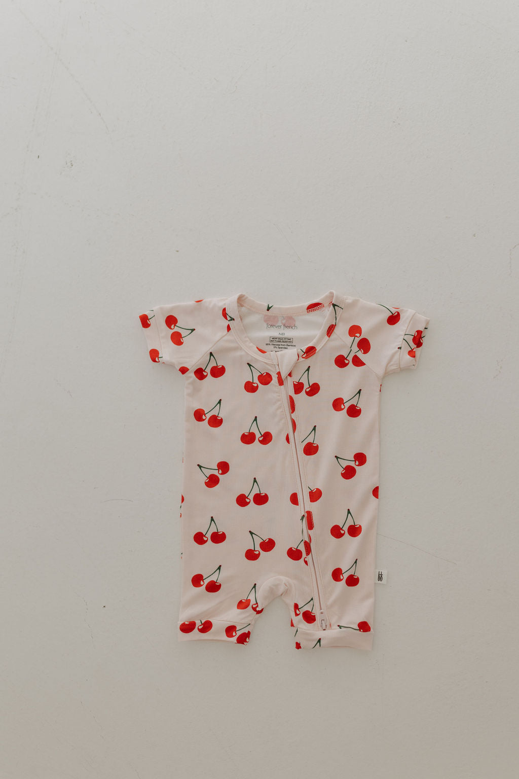 The Bamboo Zip Shortie | Cherry Orchard by forever french baby is a short-sleeved romper featuring red cherries and green stems on a light background. Made from breathable bamboo, it provides comfort for sensitive skin, while the visible seam adds subtle style.