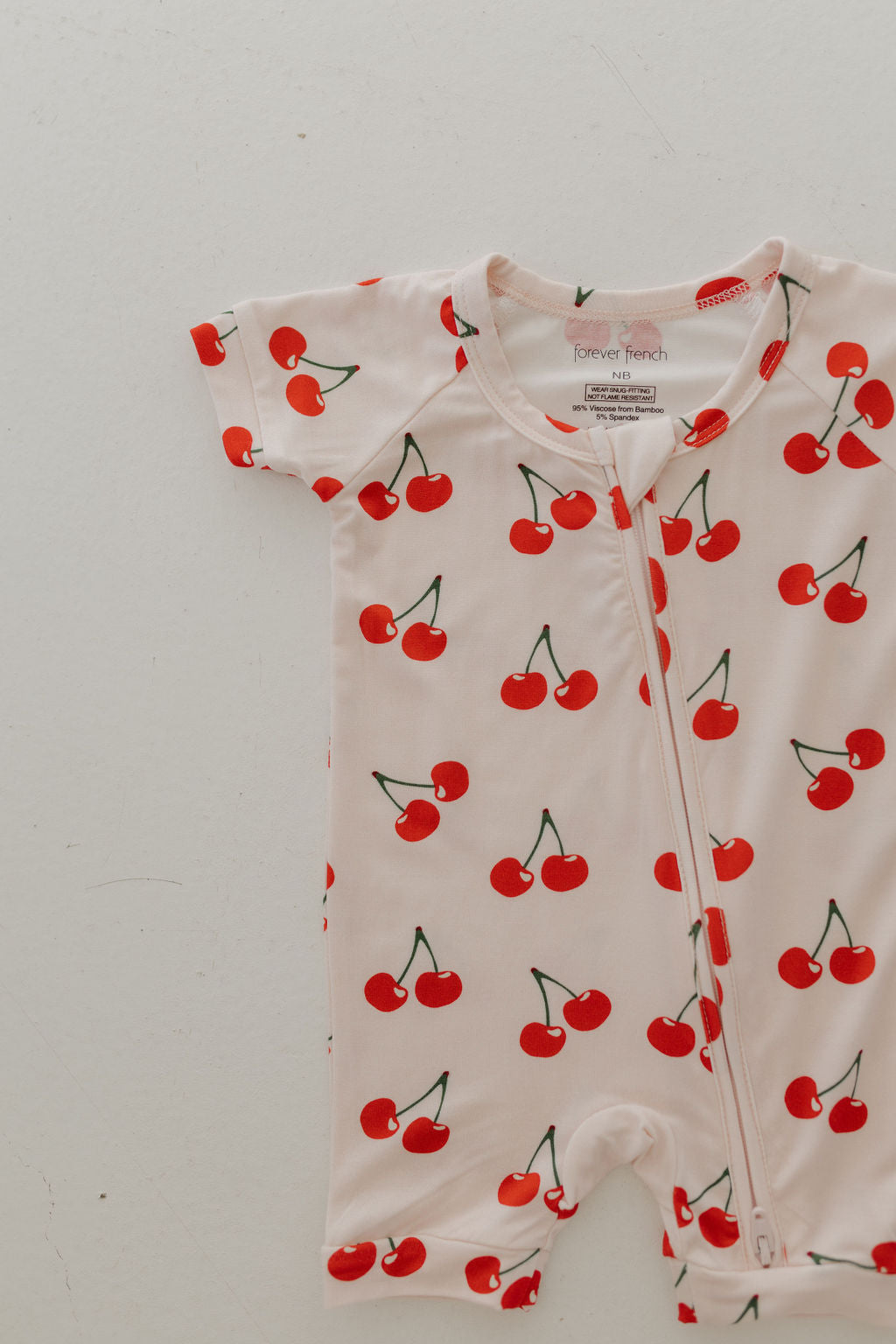The Bamboo Zip Shortie | Cherry Orchard by forever french baby is a breathable, short-sleeved romper made from hypo-allergenic fabric. It features a front zipper and vibrant red cherry print on a white background.