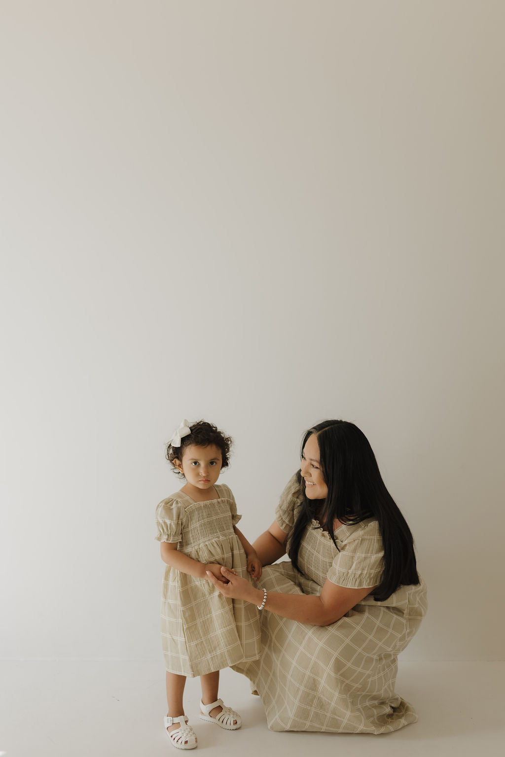 A woman kneels beside a young girl with curly hair, a white bow, and sandals. Both wear forever french baby Toddler Muslin Dress | Linen Grid—matching beige plaid 100% cotton dresses. The background is plain and light-colored.