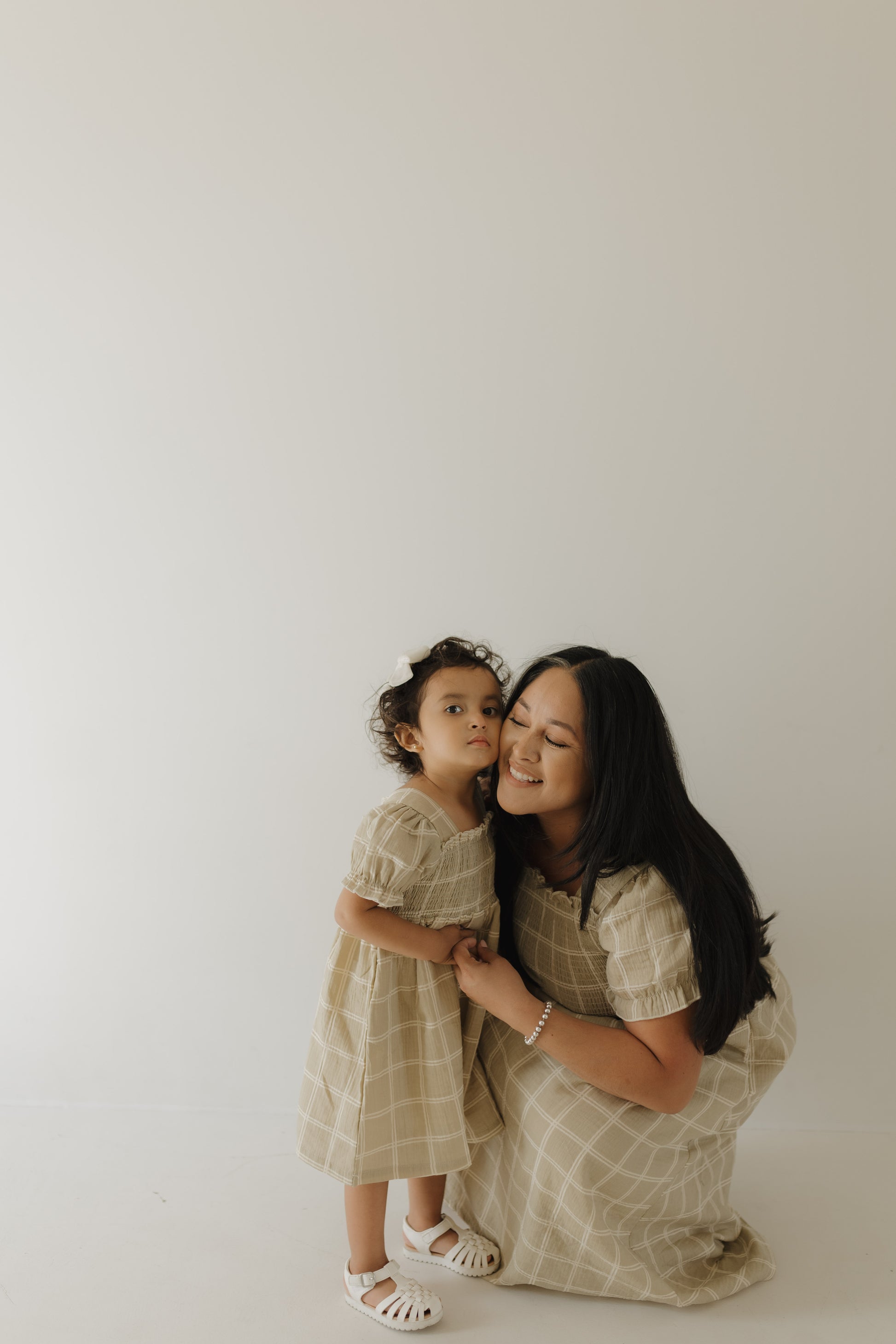 A woman kneels, smiling with eyes closed, holding hands with a young girl. Both wear forever french baby Toddler Muslin Dress | Linen Grid—100% cotton muslin. The girl's neutral gaze contrasts with the plain light background.