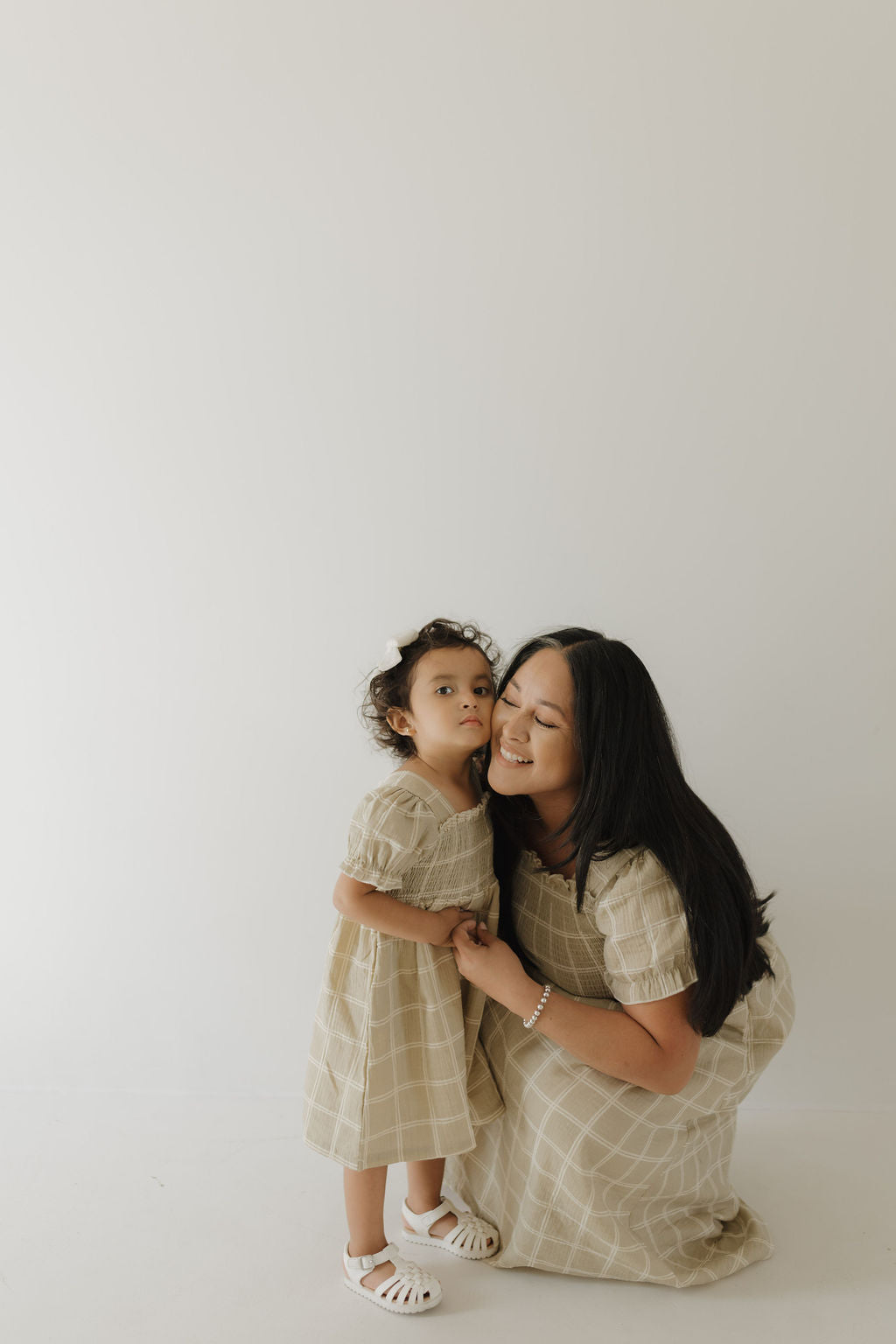 A woman kneels, smiling as she holds hands with a young girl in matching forever french baby Toddler Muslin Dress | Linen Grid outfits. Their 100% cotton dresses stand out against the plain background, highlighting their breathable charm.