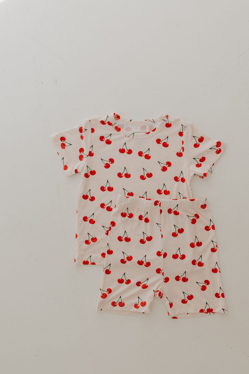 The Bamboo Short Set | Cherry Orchard by forever french baby features a children's short-sleeve shirt and shorts with a cherry pattern on a white background made from breathable, hypo-allergenic fabric. Red cherries with green stems are evenly scattered across the material, displayed flat on a white surface.