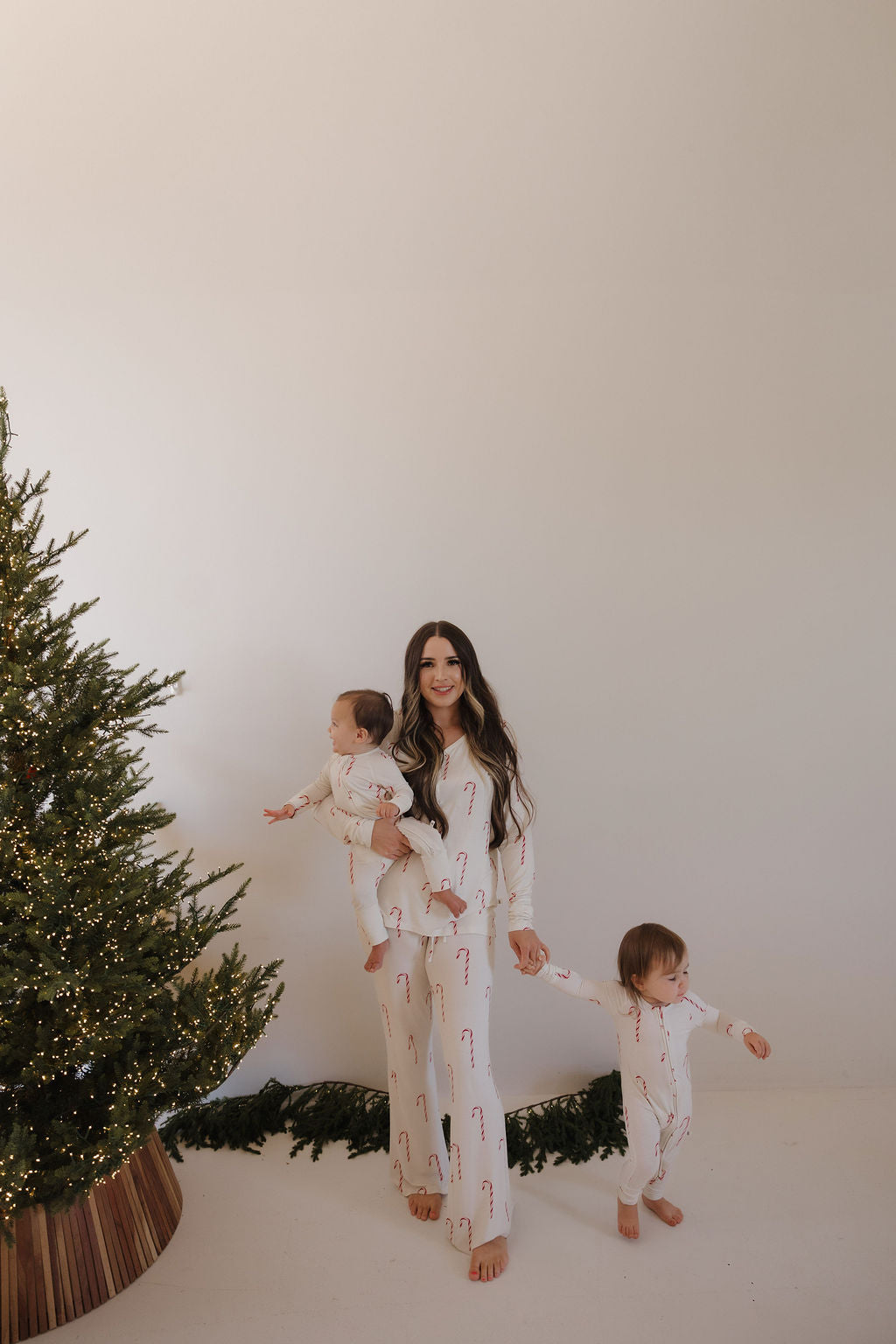 A woman in forever french baby Bamboo Zip Pajamas | Candy Cane stands by a Christmas tree, holding a baby in hypoallergenic clothing and holding hands with a toddler, surrounded by white walls and festive greenery.