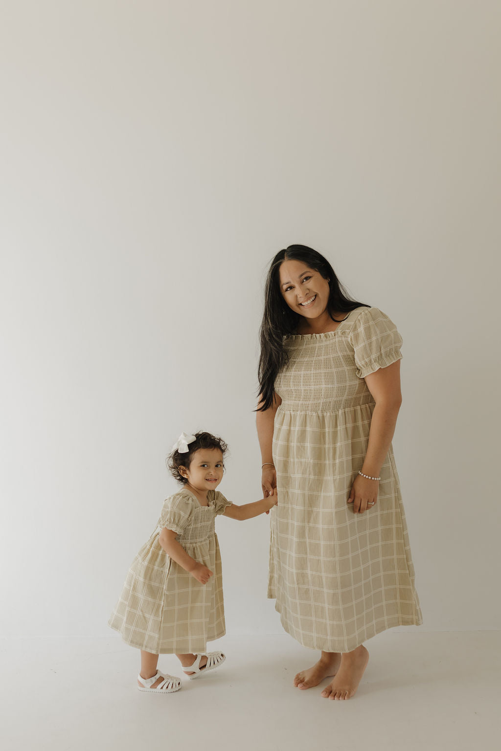A woman and a young girl with long dark hair stand barefoot, holding hands and smiling. They wear matching forever french baby Toddler Muslin Dress | Linen Grid—breathable, 100% cotton plaid muslin. The girl sports a white bow.