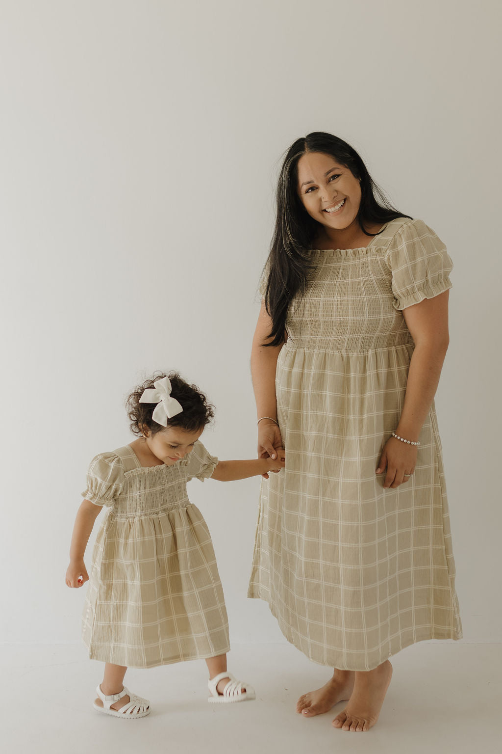 A woman and young girl, likely mother and daughter, hold hands in matching Forever French Baby "Toddler Muslin Dress | Linen Grid" outfits made from 100% cotton muslin. The girl wears a white bow with sandals; the woman is barefoot and smiling.