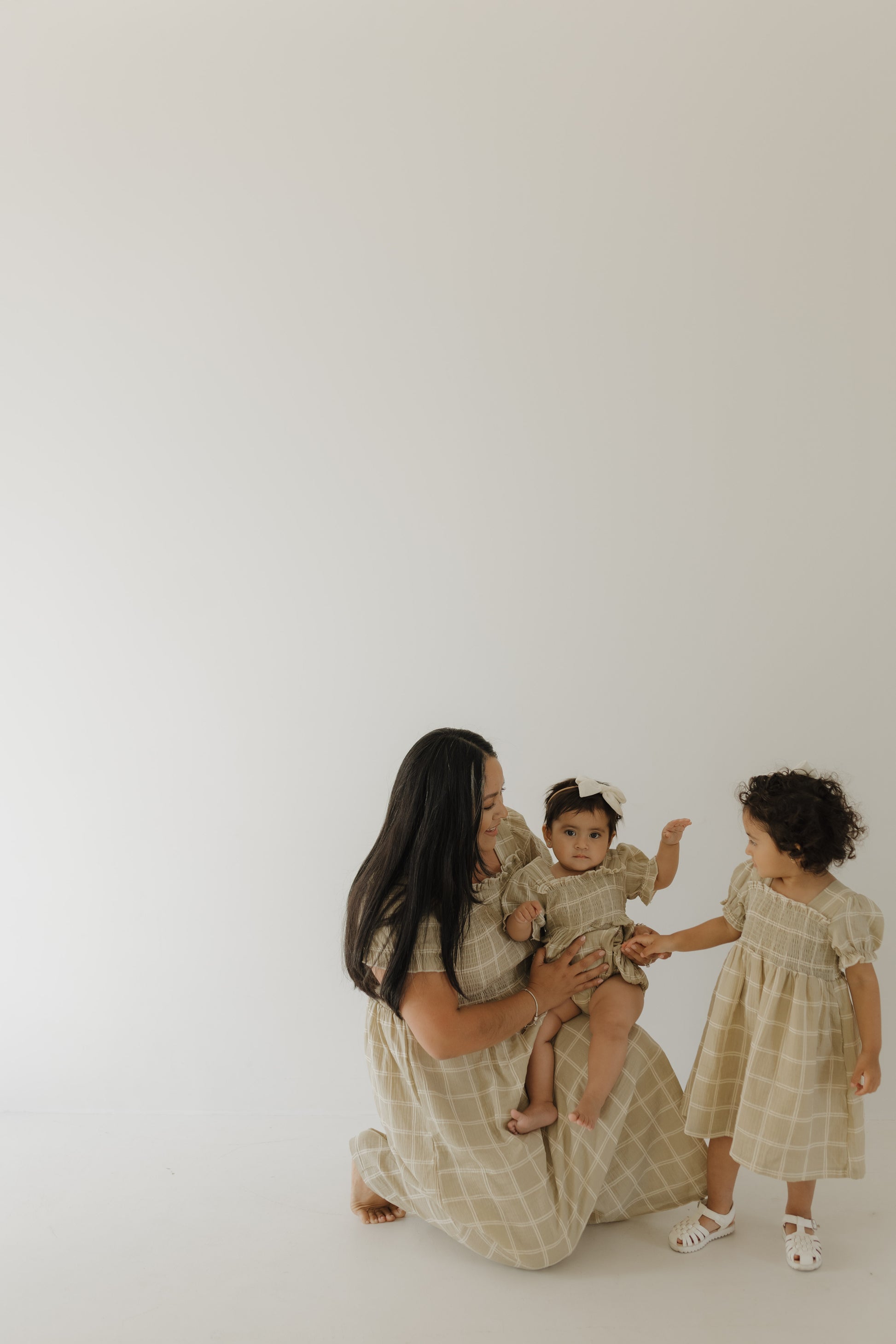 A woman kneels on the floor holding a toddler, with another child standing beside them. All wear forever french baby's Toddler Muslin Dress | Linen Grid—matching 100% cotton muslin dresses—set against a plain, light background.