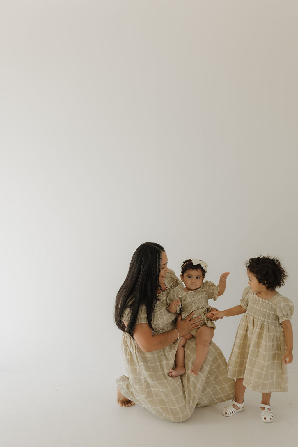 A woman kneels on a light background, holding a baby in matching muslin outfits. A young child stands beside them in the forever french baby Toddler Muslin Dress | Linen Grid, reaching toward the baby.