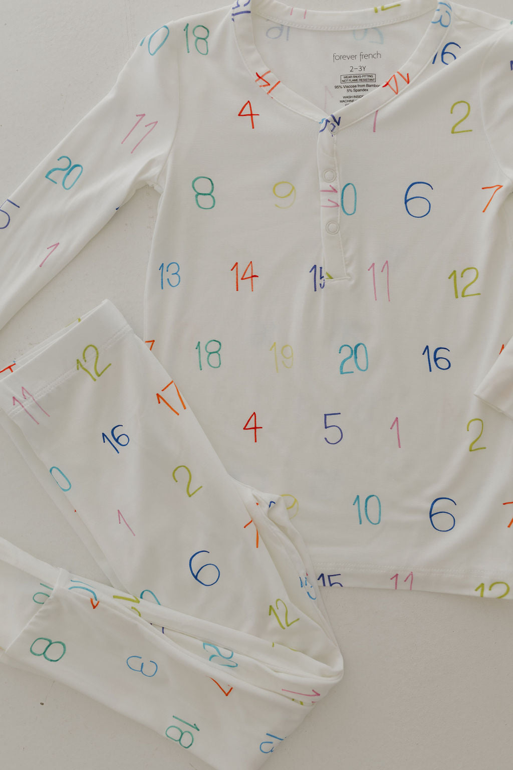 The "Charli Counts" Bamboo Two Piece Pajamas by forever french baby feature colorful numbers in different sizes on white, hypo-allergenic bamboo fabric. The set includes a number-patterned long-sleeved buttoned top and matching pants with designs like 4, 6, 14, and 20.