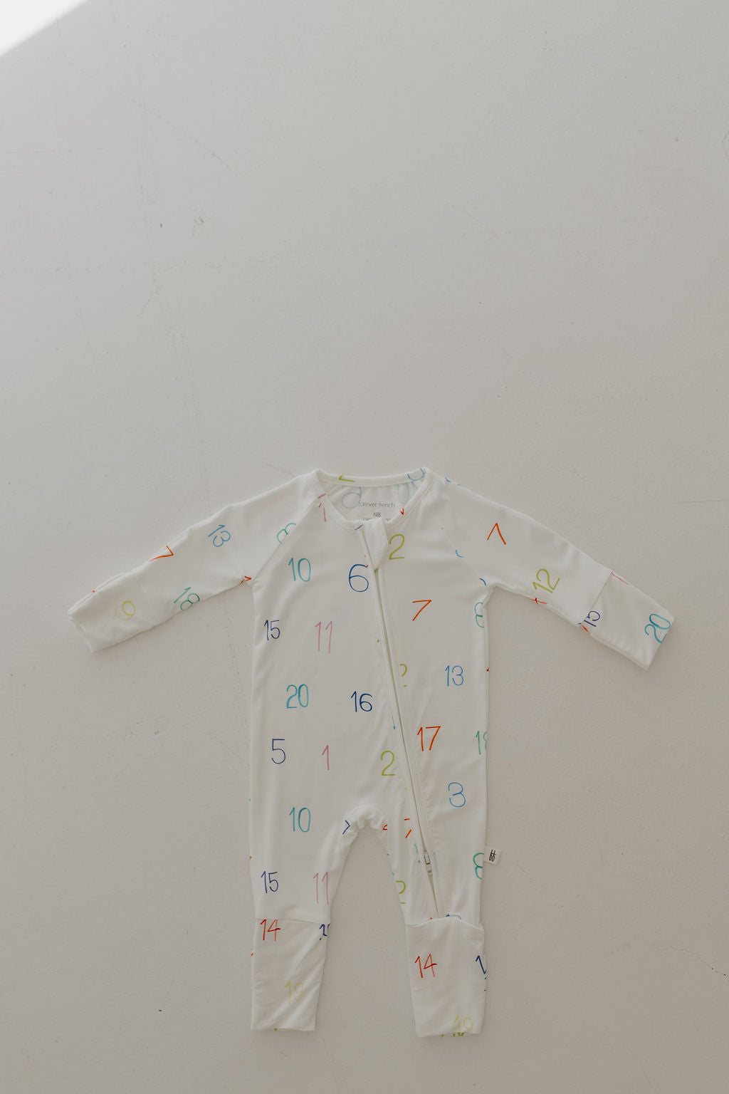 The Bamboo Zip Pajamas by forever french baby, named Charli Counts, is a white baby jumpsuit made from hypoallergenic bamboo with long sleeves and vibrant numbers scattered across the fabric. It features a front zipper and is displayed on a light surface.