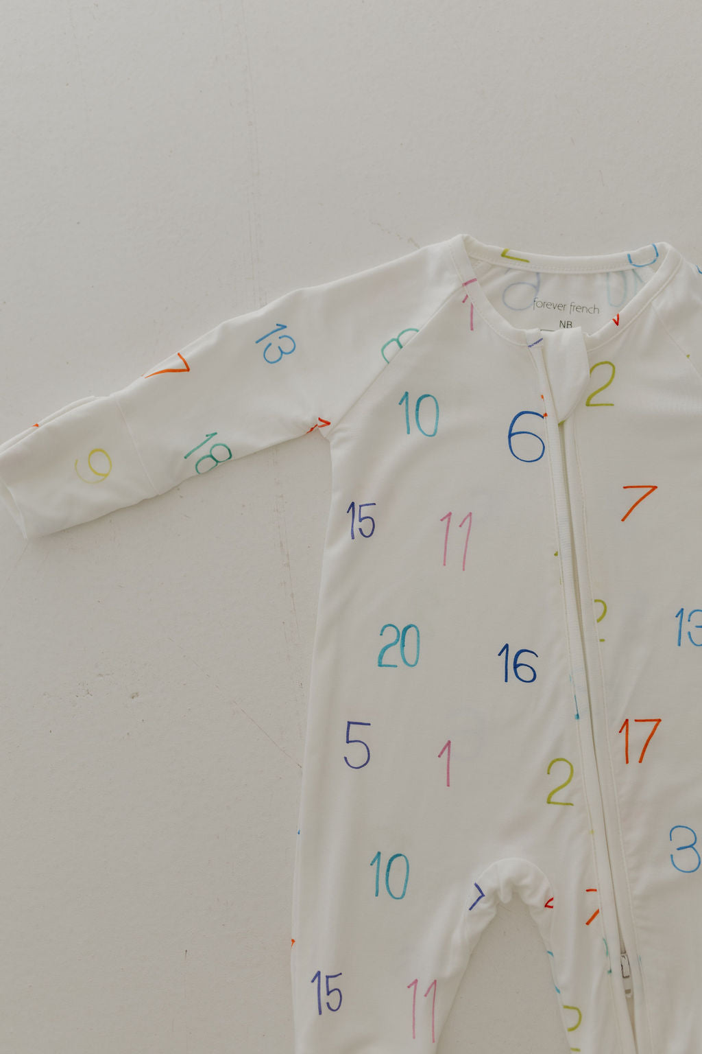 The forever french baby Bamboo Zip Pajamas | Charli Counts is a white, hypo-allergenic bamboo jumpsuit adorned with colorful numbers. It features long sleeves and a front zipper for style and comfort.