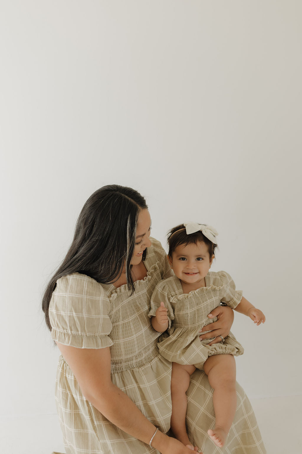 A woman and her baby girl wear matching forever french baby Women's Short Sleeve Muslin Dresses in the Linen Grid pattern, with the baby also sporting a large white bow, standing before a simple light background.
