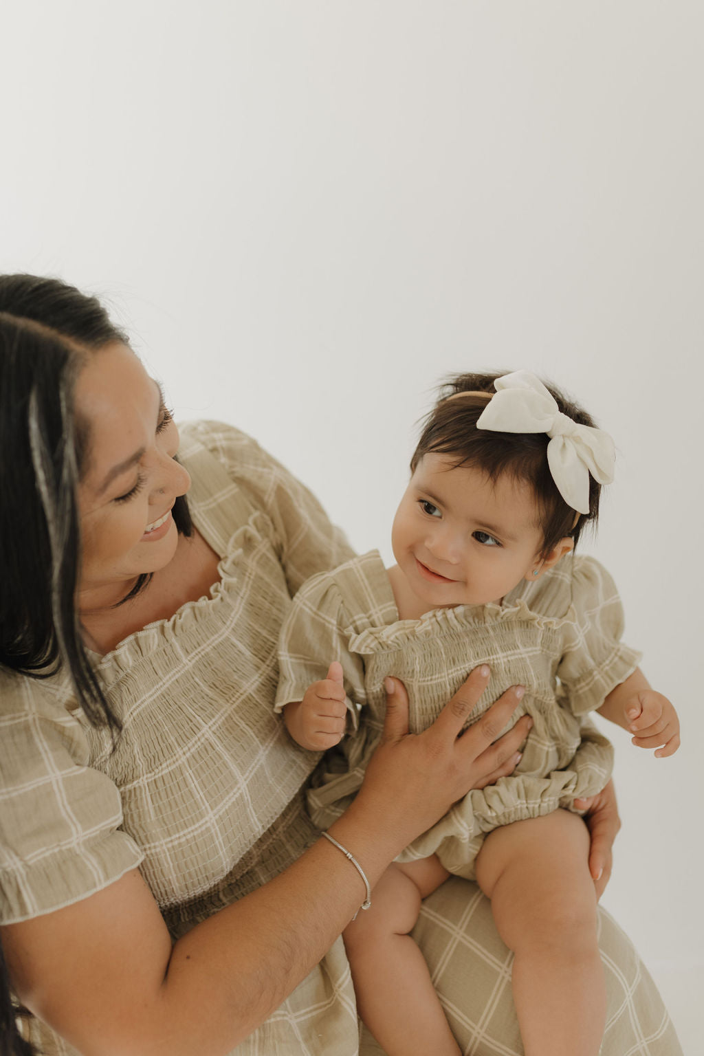A woman holds a smiling baby on her lap. Both wear matching forever french baby outfits: the Women's Short Sleeve Muslin Dress in Linen Grid for mom and a cute mini set for baby with a white bow headband, against a light background.