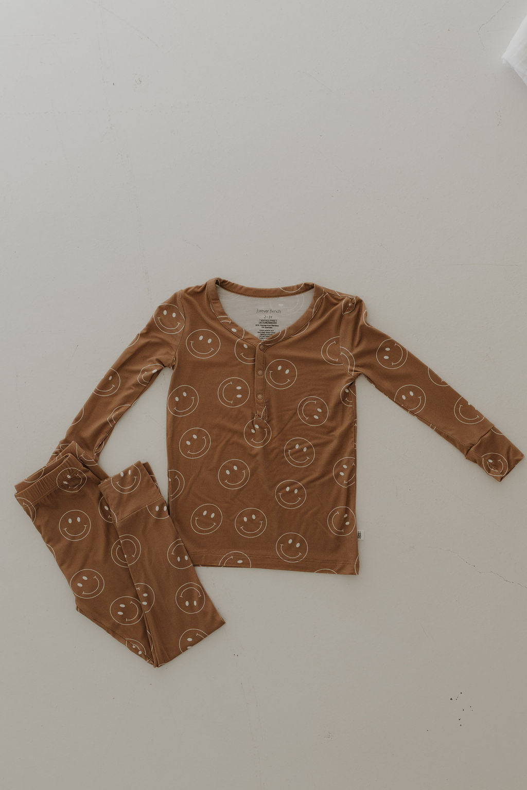 The Bamboo Two Piece Pajamas by forever french baby are displayed on a light surface, showcasing a smiling face pattern. The top has a buttoned neckline, and the pants are neatly folded beside it. Crafted from breathable fabric, this Sandstone Smile set looks soft and comfortable.