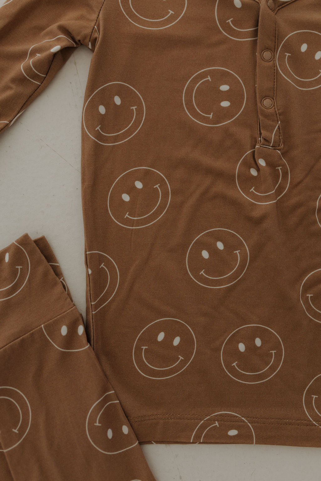 The Bamboo Two Piece Pajamas in Sandstone Smile by forever french baby are brown with white smiling faces, made from breathable, hypo-allergenic fabric. The top features long sleeves and a partially buttoned front, designed for comfort and restful nights. Image shows the shirt and part of the pants.