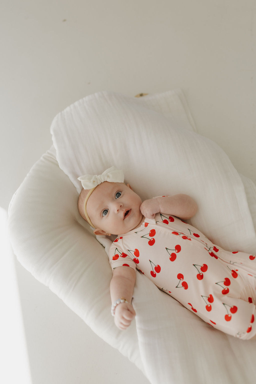 A baby lies on a soft, white cushion wearing the "Bamboo Zip Shortie | Cherry Orchard" by forever french baby. The rompers with red cherry prints and a matching bow headband are breathable, with a light, minimalistic background accentuating bamboo clothing's comfort.