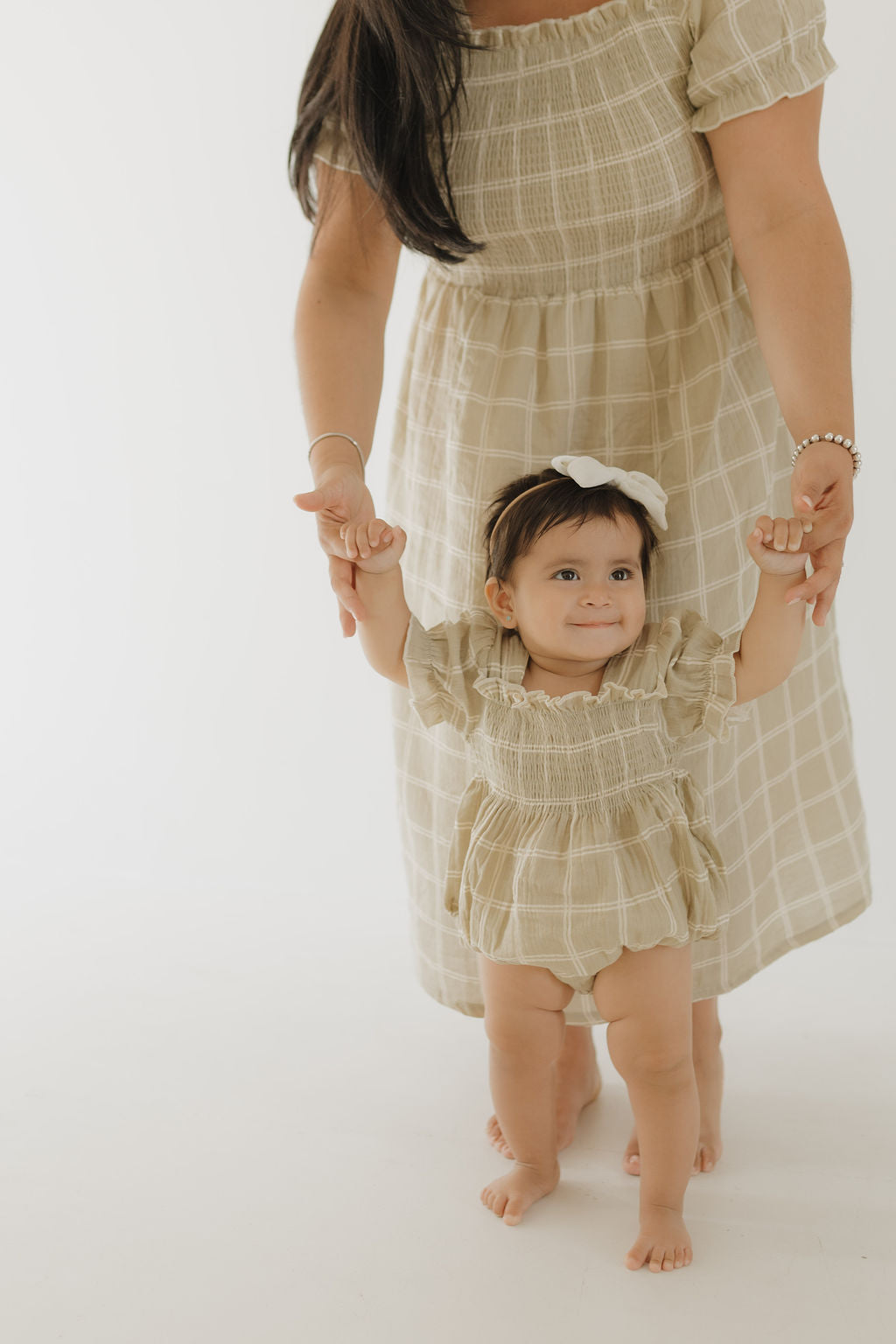 A woman in a forever french baby Women's Short Sleeve Muslin Dress | Linen Grid holds hands with a smiling toddler in matching beige plaid. Both wear 100% cotton outfits; the child sports a white hair bow. The background is plain and light.