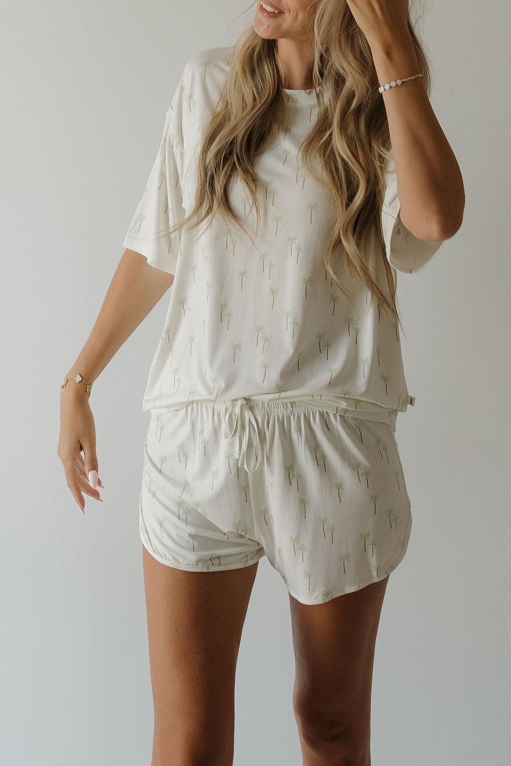 A woman models the forever french baby Women’s Bamboo Short Set | Palm—a cream-colored, short-sleeve and shorts pajama set adorned with green palm tree prints—against a plain light background. Her long hair falls in loose waves, highlighting the breathable, hypo-allergenic bamboo fabric that ensures comfort and effortless style throughout the night.
