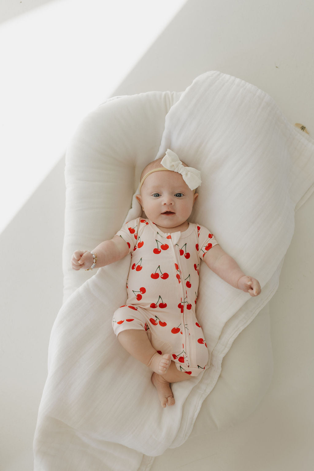 A baby in a "Bamboo Zip Shortie | Cherry Orchard" by forever french baby, adorned with a large white bow, lies on a white cushion. Sunlight casts soft shadows as they gaze upward with outstretched arms, surrounded by breathable serenity in hypo-allergenic fashion.
