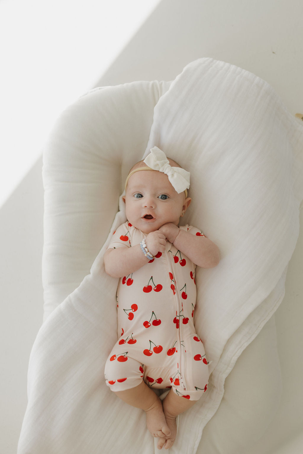 A baby lounges on a white cushion, dressed in the Bamboo Zip Shortie | Cherry Orchard from forever french baby. The light pink onesie features red cherry patterns, complemented by a white bow headband and bracelet, as the baby gazes thoughtfully in soft lighting.