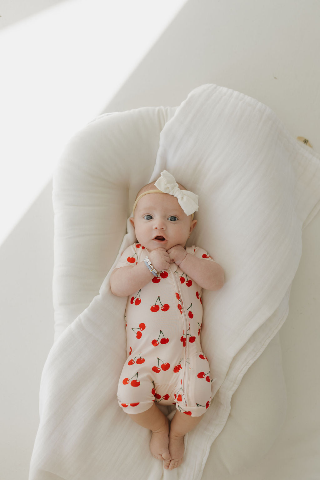 A baby lies on the soft, white surface of a blanket designed for delicate skin, wearing a hypo-allergenic Bamboo Zip Shortie in Cherry Orchard by forever french baby and a white headband with a bow. A beaded bracelet adorns their wrist as they gaze upward.