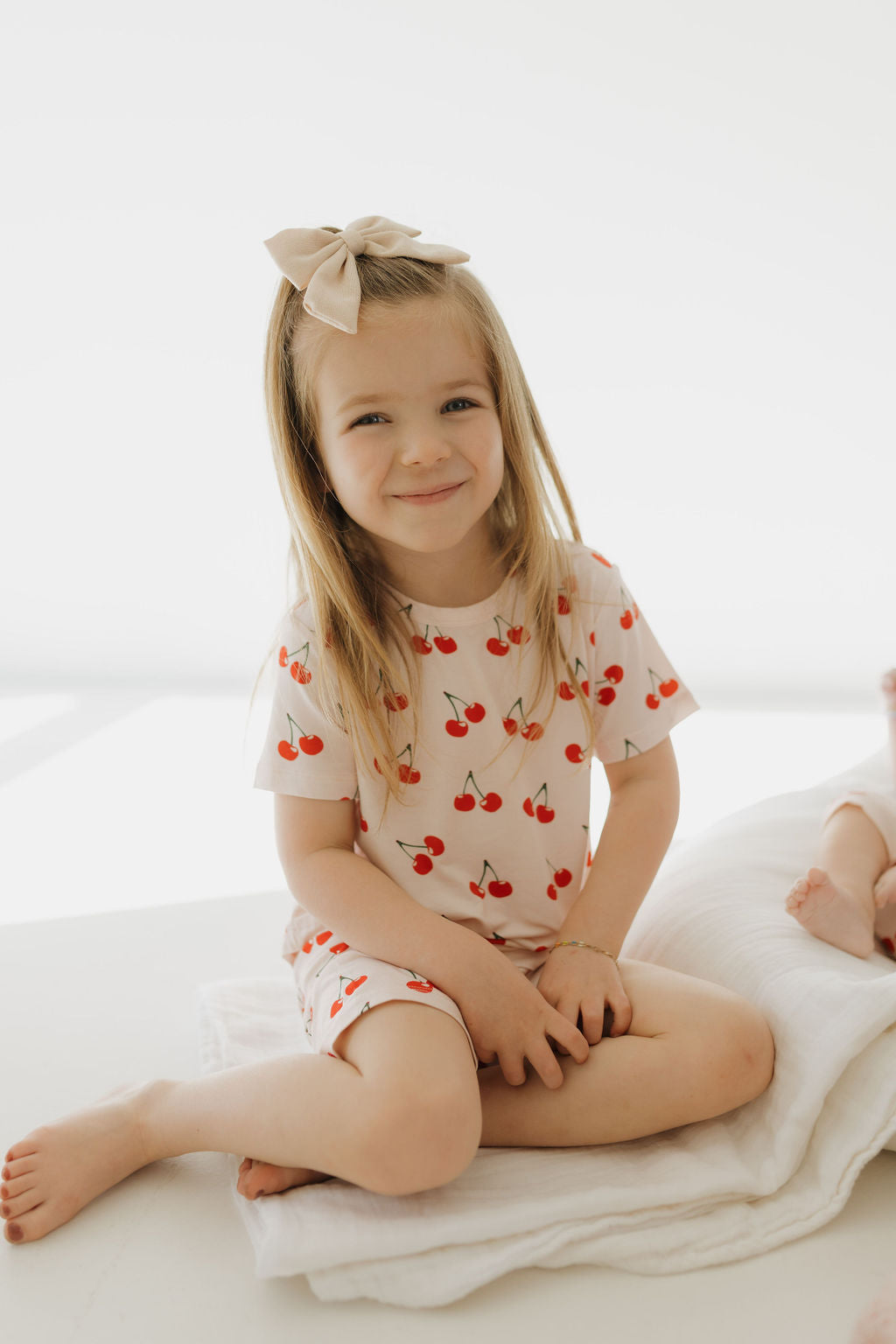 A young girl with long blonde hair and a bow sits cross-legged on a bed, smiling. She wears the "Bamboo Short Set | Cherry Orchard" by forever french baby, offering breathable comfort. The background is bright and softly lit.