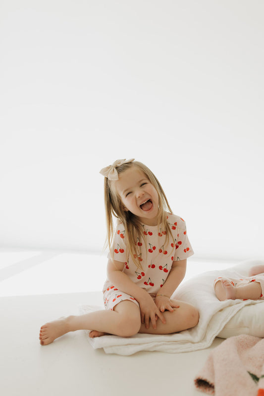 A young girl with long hair and a bow sits cross-legged on a white surface, laughing. She’s wearing the Bamboo Short Set | Cherry Orchard by forever french baby. Nearby lies a soft blanket. The scene is bright and minimalistic.
