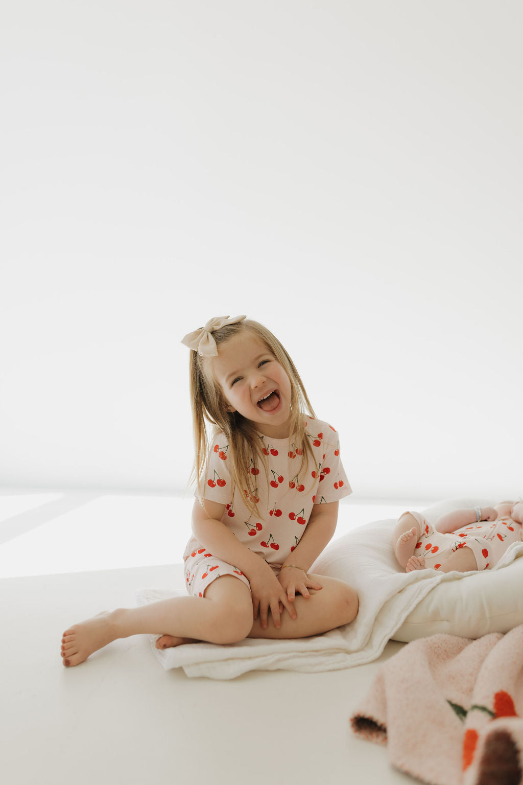 A young girl with long hair and a bow, wearing the Bamboo Short Set | Cherry Orchard from forever french baby, sits cross-legged on a white cushion, smiling brightly. A patterned blanket and part of another child are visible on the right against a bright white background.