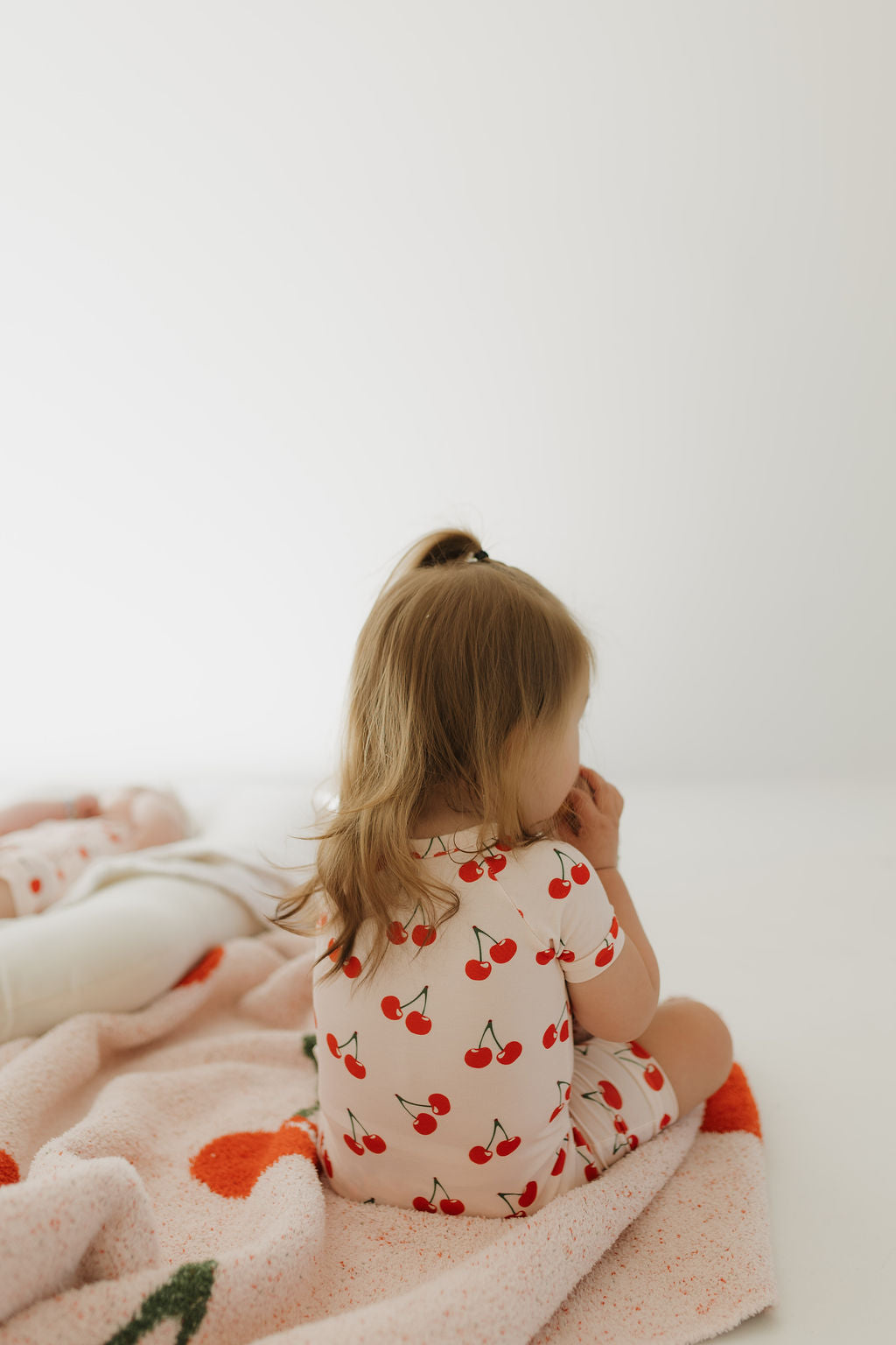 A child with light brown hair in a ponytail sits on a bed, wearing forever french baby's "Bamboo Zip Shortie | Cherry Orchard" hypo-allergenic pajamas. The playful bedding features large orange circles, and the softly lit room adds serenity as the child faces away from the camera.