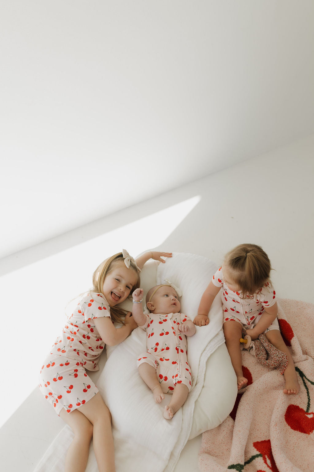 On a light floor, two toddlers and a baby, all dressed in forever french baby's Bamboo Short Set | Cherry Orchard with white fabric adorned with red cherries, lie together. The toddlers smile while the baby rests comfortably on a plush white cushion as sunlight streams beautifully.