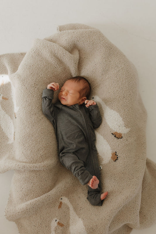 A newborn baby in forever french baby Bamboo Zip Pajamas | Shadow, a hypo-allergenic dark gray outfit, lies peacefully on a soft beige blanket with white bird patterns, eyes closed and one arm raised.