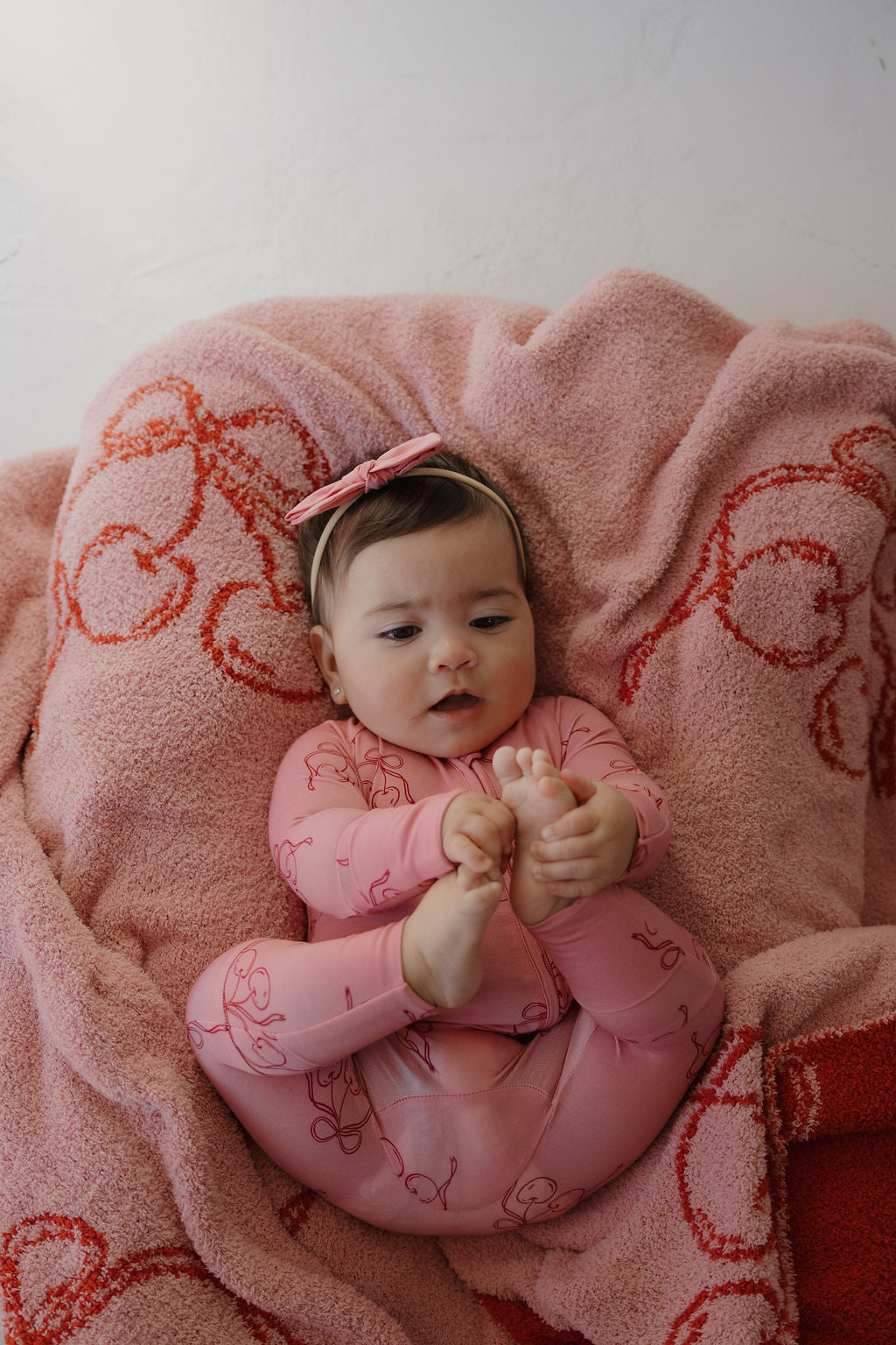 A baby in Minty Cherries' Bamboo Zip Pajamas | Iconic Cherry—featuring a pink outfit with red line patterns—lies on a matching pink blanket, holding their feet and looking up for a coordinated, cozy look.