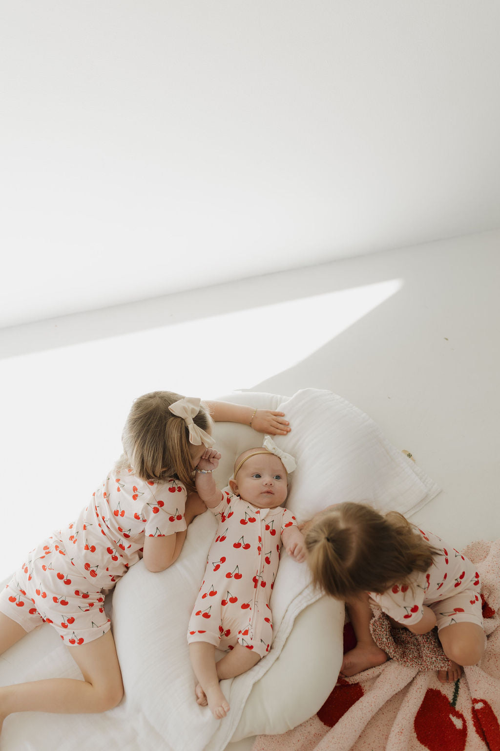 Three young children in hypo-allergenic Bamboo Zip Shortie by forever french baby, Cherry Orchard design, play on white pillows and a pink blanket. Two sit engaged beside the infant lying on its back looking up, creating a serene and playful scene in soft lighting.