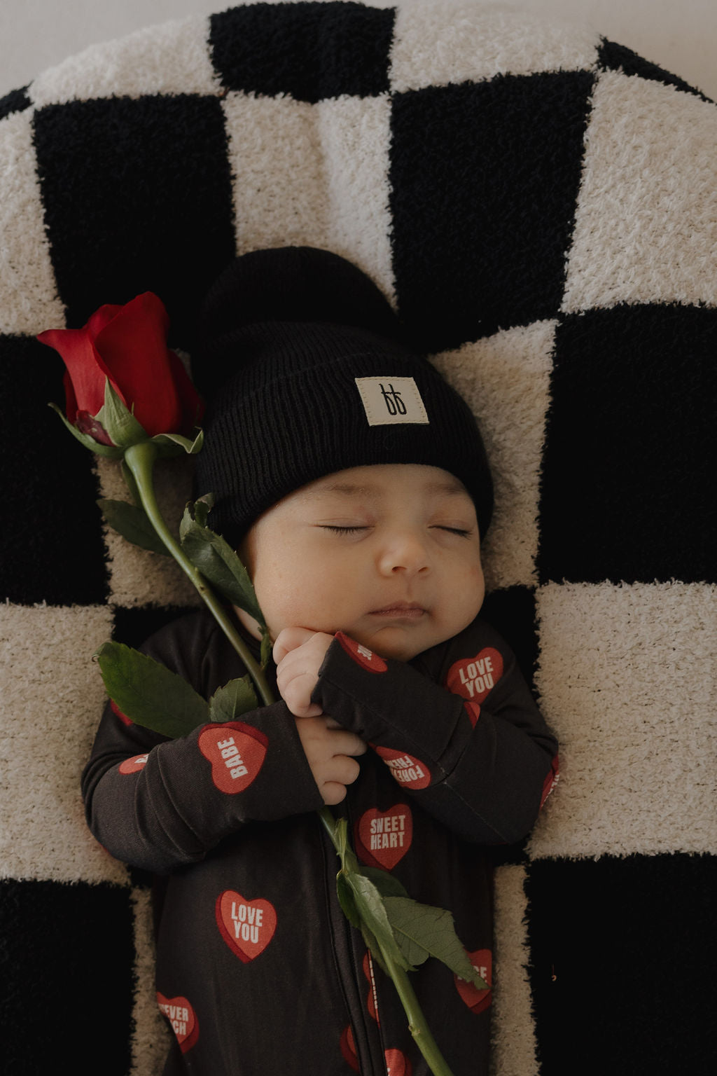 A sleeping baby in forever french baby's Bamboo Zip Pajamas | Love Notes – Charcoal holds a red rose while lying on a black-and-white checkered blanket, wearing heart-patterned pajamas and a matching black beanie.