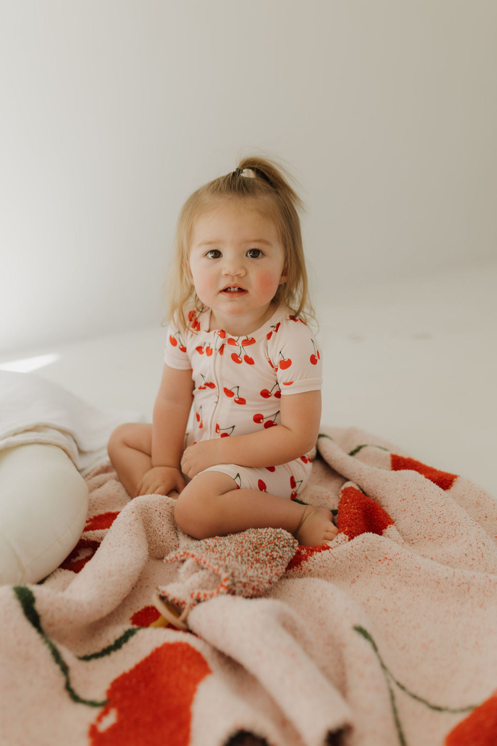 A toddler with a small ponytail sits on a bed, wearing the Bamboo Zip Shortie | Cherry Orchard by Forever French Baby. They're on a pink and red blanket, with a plush toy nearby. The soft, light background creates a serene scene.