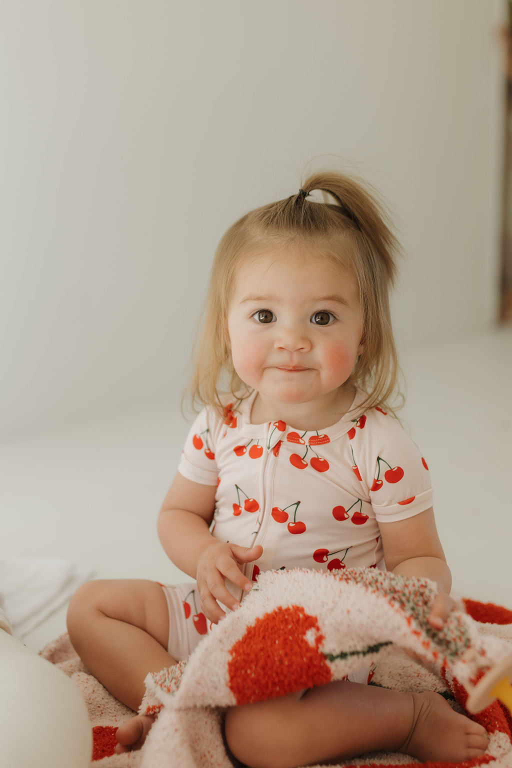 A toddler with light brown hair in a small ponytail sits on the floor, wearing a Bamboo Zip Shortie from forever french baby's Cherry Orchard collection. She clutches a textured blanket, while the softly blurred background creates a warm and cozy atmosphere.