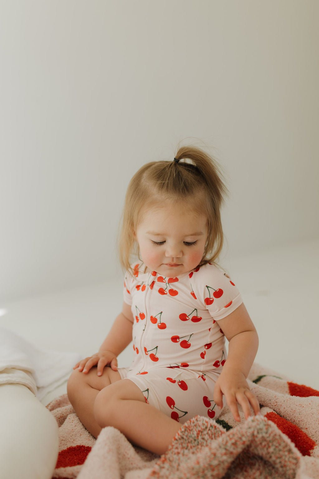 A toddler with light brown hair sits on a blanket, dressed in Forever French Baby's Bamboo Zip Shortie in Cherry Orchard, featuring breathable fabric and cherry designs. The child gazes calmly at the blanket against a softly lit, neutral background.