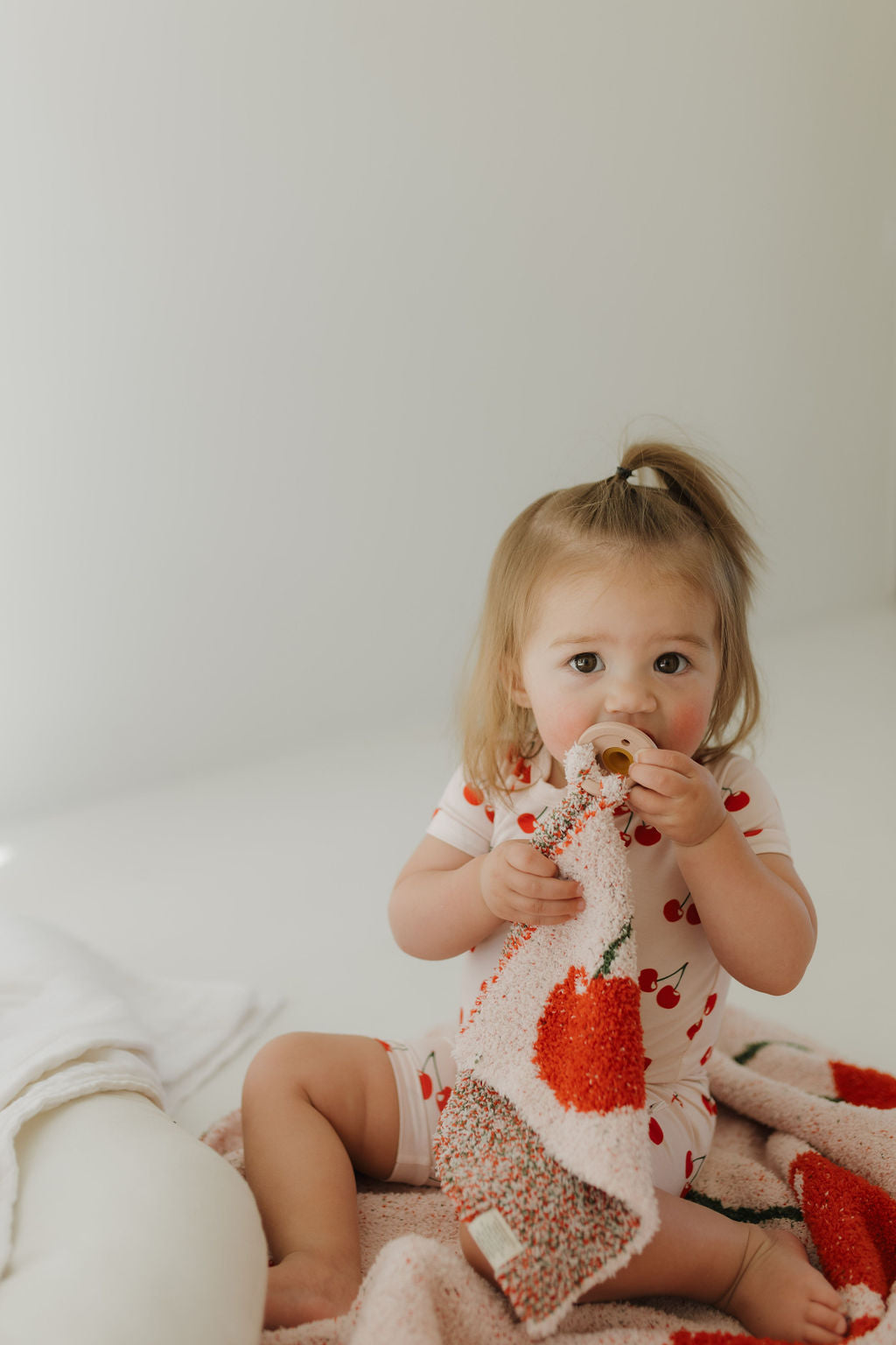 A toddler with light brown hair sits on a soft surface, wearing the "Bamboo Zip Shortie | Cherry Orchard" by forever french baby. She holds a pacifier and a pink blanket with an orange strawberry design. The plain, light-colored wall enhances focus on her adorable outfit.
