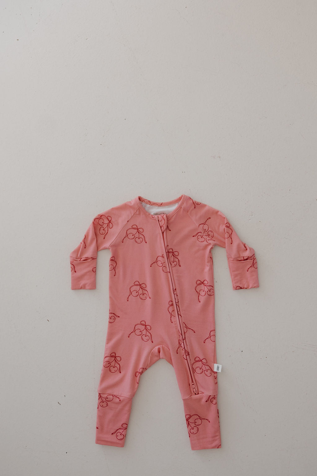 The Minty Cherries Bamboo Zip Pajamas in Iconic Cherry feature breathable long sleeves, footed design, a simple red cherry pattern, and a diagonal zipper from neck to ankle, shown laid flat on a light surface.