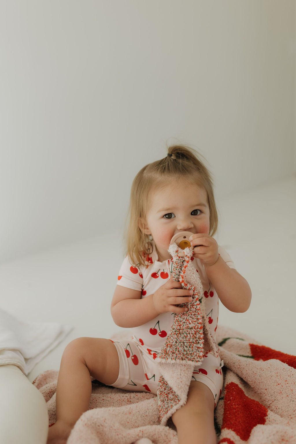 A toddler with light brown hair sits on a bed, wearing the "Bamboo Zip Shortie | Cherry Orchard" romper from forever french baby, adorned with red cherries. The child holds a pacifier and a textured pink blanket against a neutral, minimalist background.