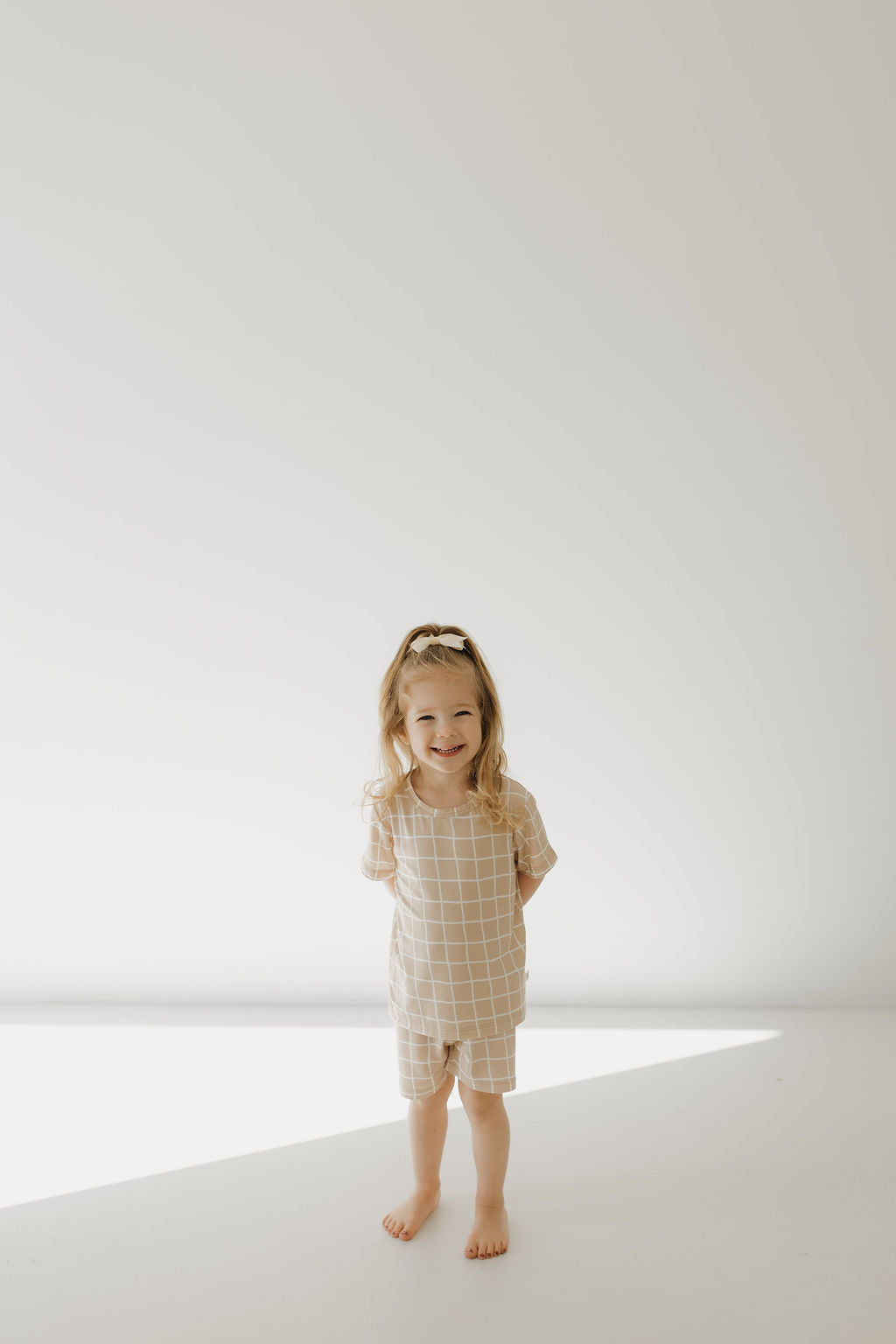 A young child with long hair, barefoot in a bright, minimalist space, wears the Bamboo Short Set | Grid from forever french baby. The hypo-allergenic pajamas in beige checkered bamboo enhance their joyful smile as they stand with hands behind their back, softly lit by natural light.