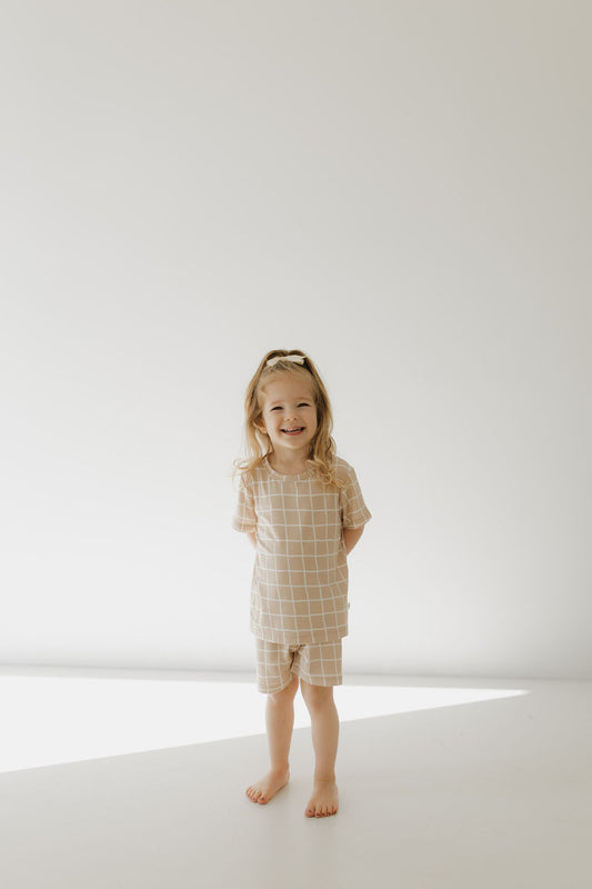 A young child with long hair smiles while standing barefoot in a well-lit room, dressed in the Bamboo Short Set | Grid by forever french baby. The tan pajamas feature a white grid pattern, offering breathable comfort and hypo-allergenic benefits for sensitive skin against a plain white wall with soft lighting.