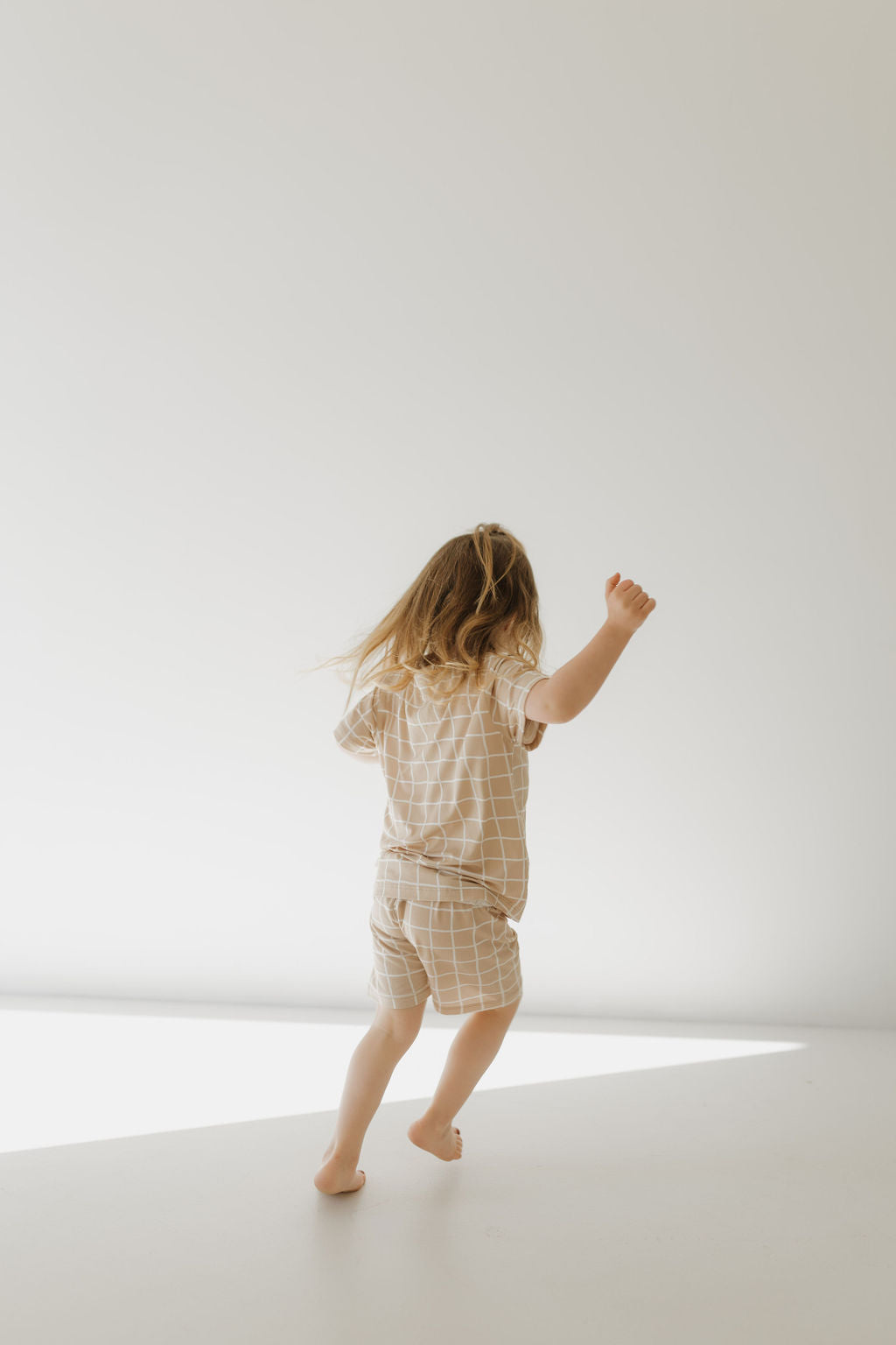 A child with long hair energetically walks barefoot in a sunlit, minimalistic space, wearing a breathable Bamboo Short Set | Grid by forever french baby. Their arm is raised against a plain and softly lit background.