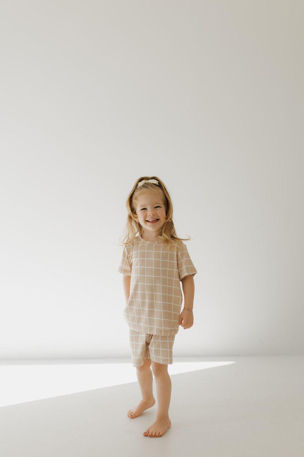 A child stands barefoot against a light background, smiling widely in forever french baby's hypo-allergenic Bamboo Short Set | Grid. The matching beige outfit with a white grid pattern complements their long, flowing hair in a minimally set scene enhanced by soft, natural lighting.