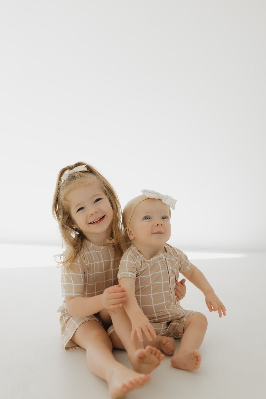 Two young children sit on a light background in matching "Bamboo Zip Shortie | Grid" outfits by forever french baby. Both wear white headbands. The older child smiles gently while holding the curious younger one who gazes upward.