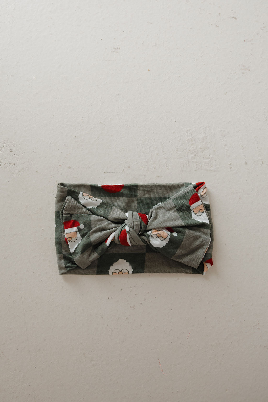 The Bamboo Head Wrap | Santa Checkerboard by forever french baby features green plaid with Santa faces in red hats and a knotted center, laid flat on a light surface. Perfect festive accessory for little ones.